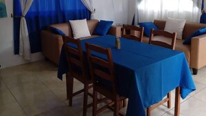 A dining area with a table set for four, surrounded by wooden chairs. There are two sofas with blue and white cushions, and a wall-mounted fan.