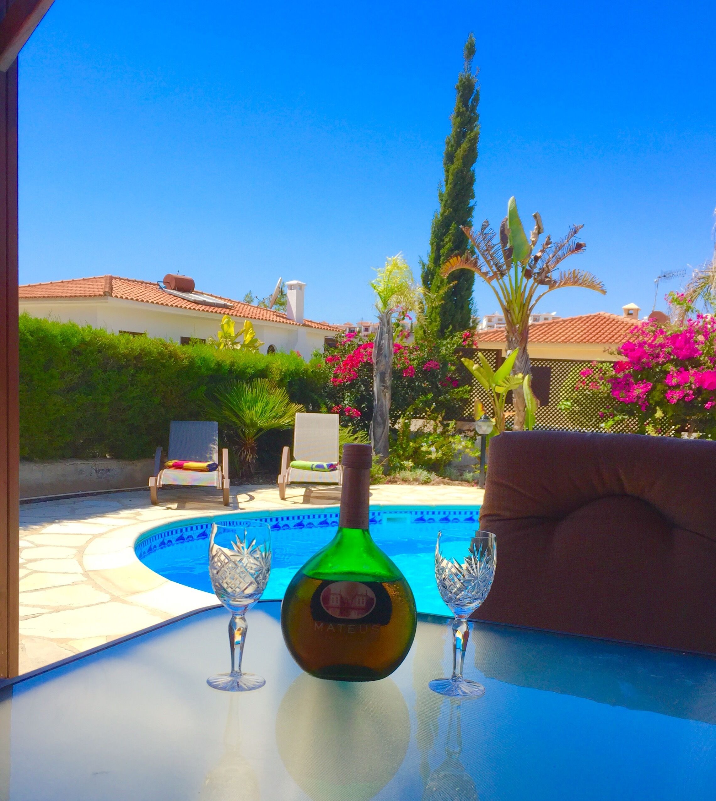 A pool table with a bottle and two glasses, surrounded by outdoor furniture and a pool.
