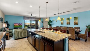 A spacious kitchen with a central island, a dining area with wicker chairs, and a living room with a sofa and a TV.