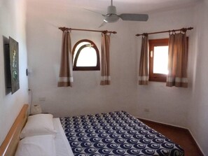 A bedroom with a bed, a ceiling fan, two windows with curtains, and a wall-mounted picture. - House with garden (Villasimius)