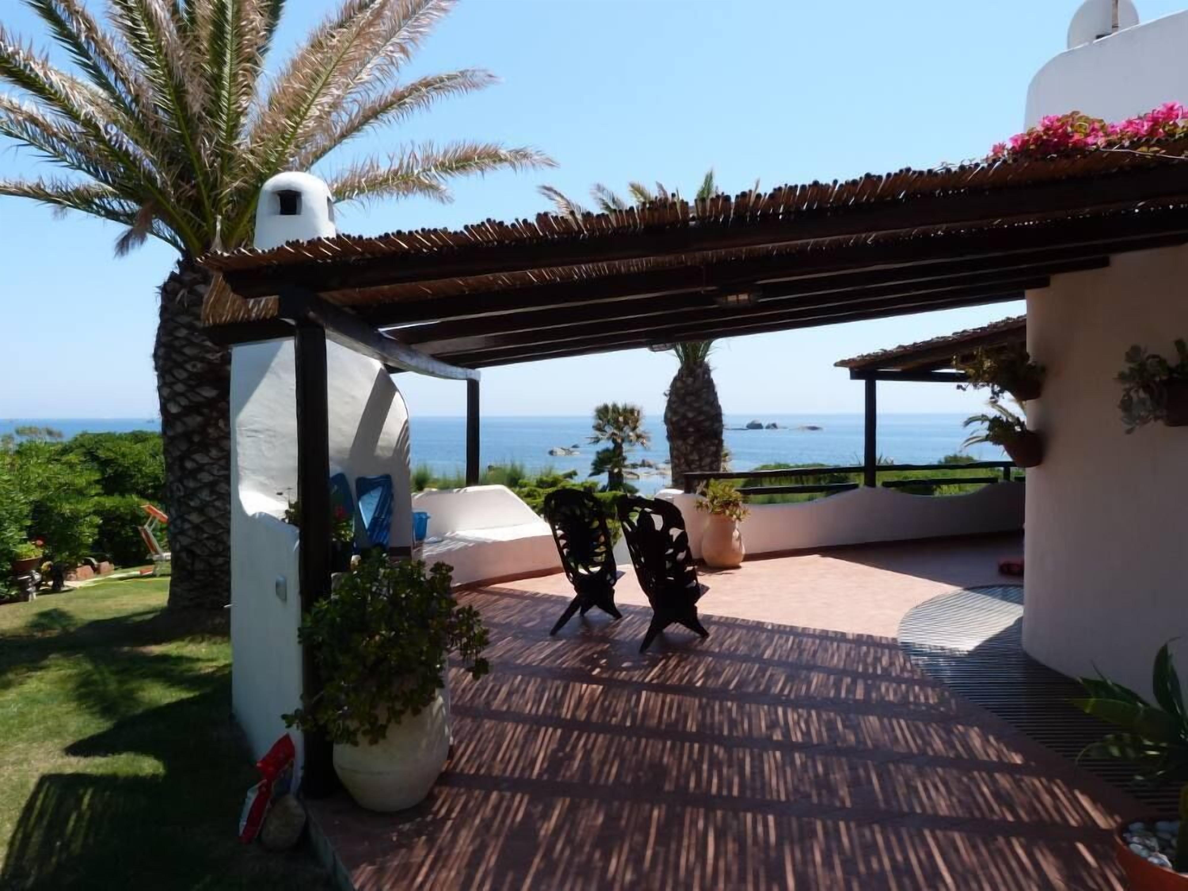 A covered outdoor seating area with a view of the sea, a white building, and a palm tree.