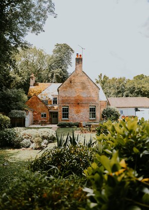 Une maison de campagne en briques, dotée d’une cheminée imposante, entourée d’une végétation luxuriante et d’un jardin bien entretenu.