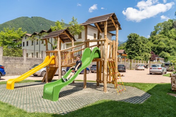 A playground with slides and climbing structures, surrounded by a paved area and parked cars. - Residence Toli Pool with outdoor heated Pool 30° (Ledro)