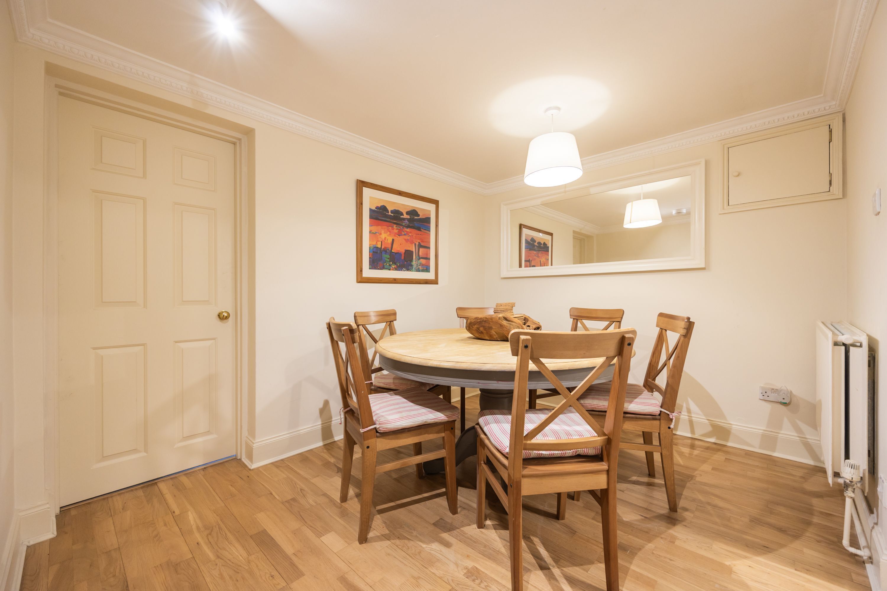 A dining room with a round table, six wooden chairs, a painting on the wall, and a mirror above the table.