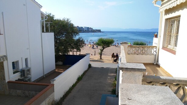 Ein schmaler Gang mit Blick auf einen Strand und das Meer. An beiden Seiten des Ganges befinden sich Gebäude, und in der Mitte steht ein kleiner Baum.