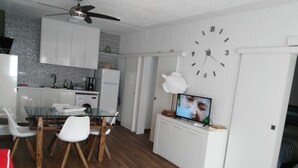 Eine kompakte Küche mit weißen Schränken, eine Essecke mit einem Glastisch und weißen Stühlen, eine Uhr an der Wand und ein Fernseher auf einem Schrank.
