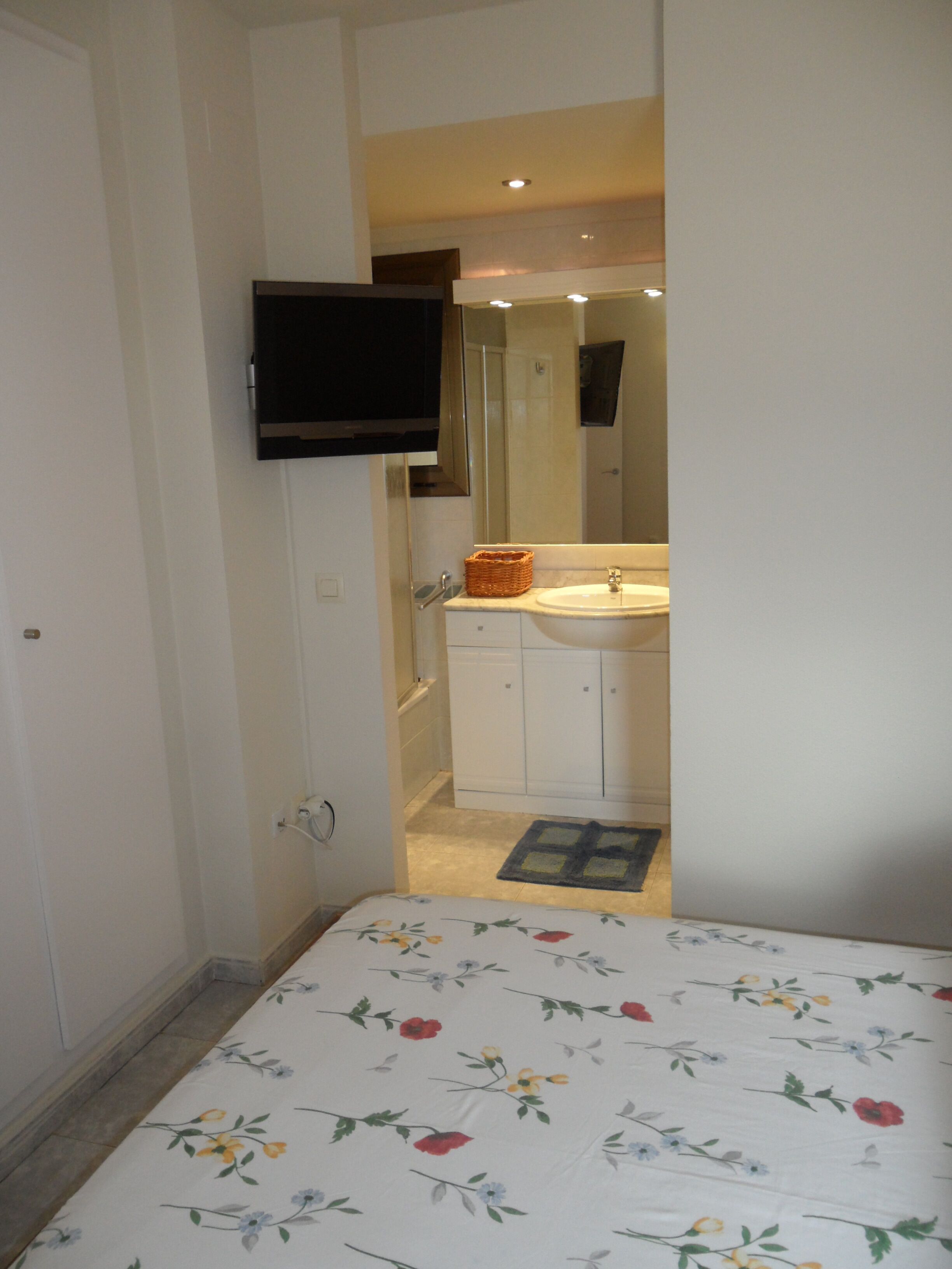 A bedroom with a bed, a bedside table, a nightstand, a wall-mounted TV, and a bathroom visible through an open door.