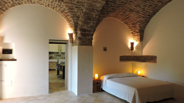 A bedroom with a stone ceiling, a bed, a nightstand with a lamp, and a view into the kitchen.