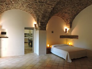 Een slaapkamer met een stenen plafond, een bed, een nachtkastje met een lamp en uitzicht op de keuken.