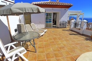 Um terraço com cadeiras de lounge brancas e uma mesa, um guarda-sol e vista para o mar.