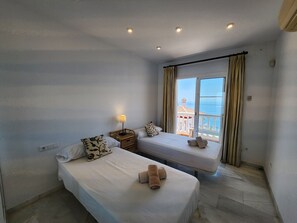 Quarto de hotel com duas camas, uma mesa de cabeceira com abajur e vista para o mar através de uma janela grande.