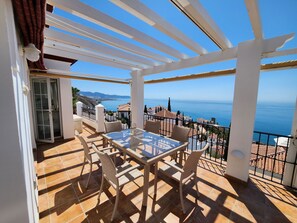 Varanda com mesa de vidro e cadeiras de vime, com vista para a costa, casas e o mar.