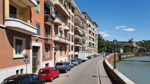Una hilera de edificios residenciales de varios pisos con balcones, coches aparcados a lo largo de la calle y un río al fondo.