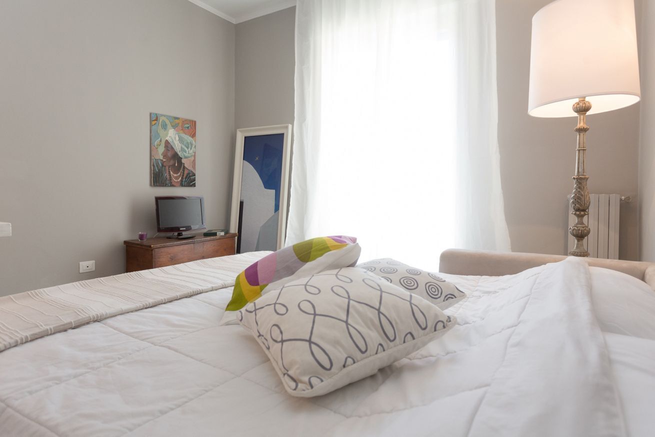 Een net opgemaakt bed met witte beddengoed en decoratieve kussens, een nachtkastje met een lamp, een televisie en een schilderij aan de muur.