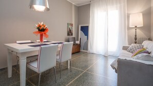 Una sala de estar moderna con comedor, que incluye una mesa blanca, sillas, una lámpara colgante grande, un sofá y un ventanal con cortinas.