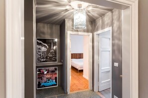 Ein Flur mit einer Chandelüster, ein Bett im angrenzenden Zimmer und gerahmte Bilder an der Wand.