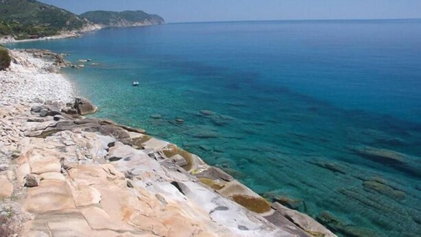 A coastal landscape with clear blue waters, rocky terrain, and distant hills.