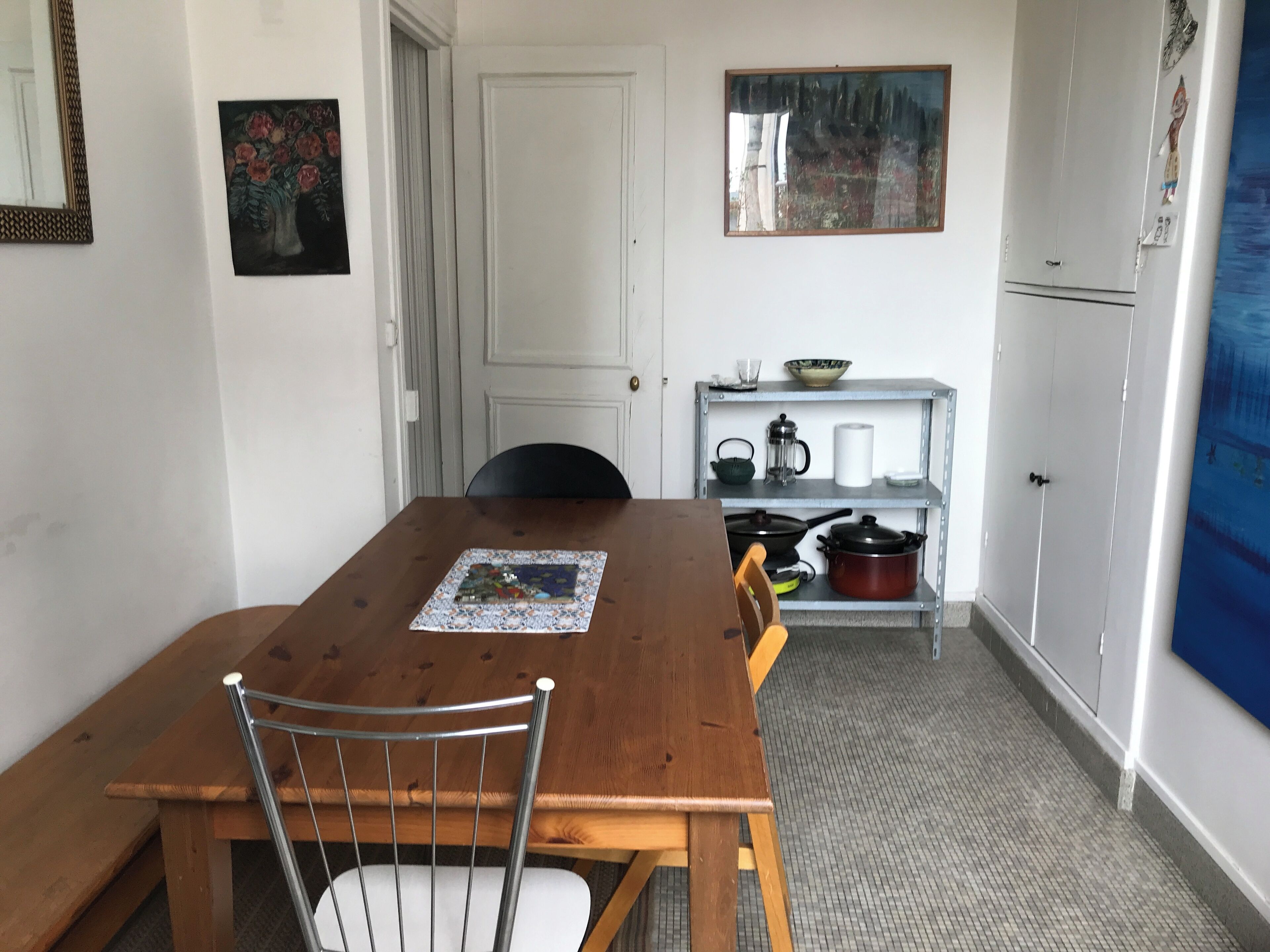 Un comedor con una mesa de madera, sillas, un cuadro y una zona de cocina con estantes y electrodomésticos.