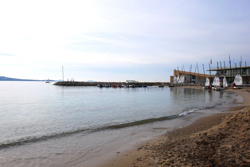 Escena costera con playa arenosa, aguas tranquilas y un edificio con varios veleros amarrados.