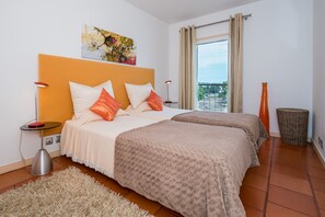 A hotel room with a large bed, a small side table, a floor lamp, a waste basket, and a window with curtains.