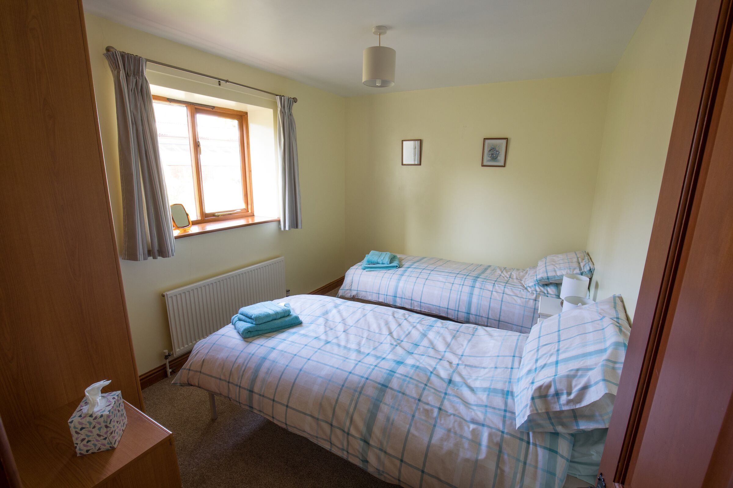 A bedroom with two beds, a window with curtains, a radiator, and a bedside table with a tissue box.