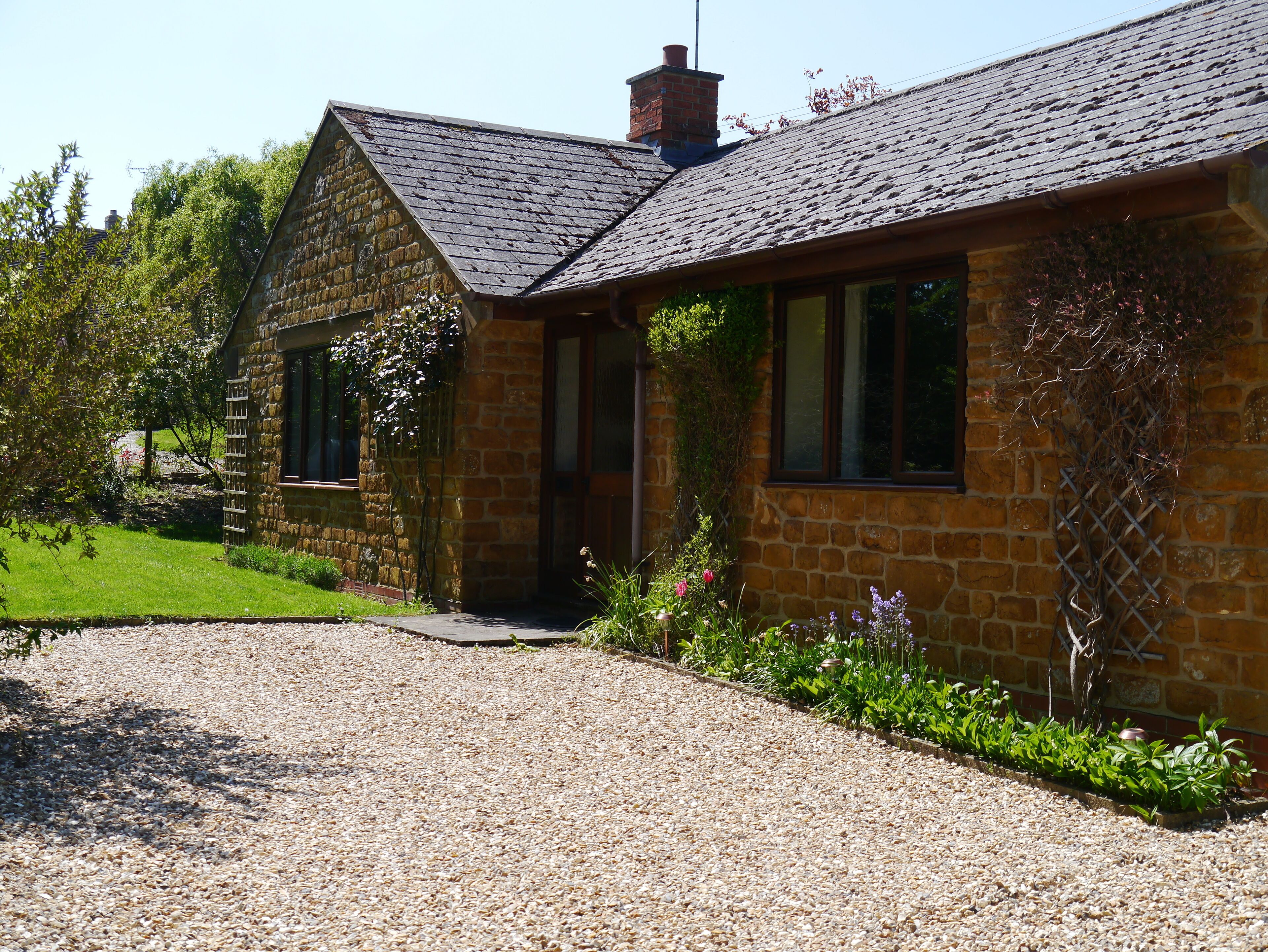 A stone house with a gravel driveway, a wooden fence, and a garden with various plants.