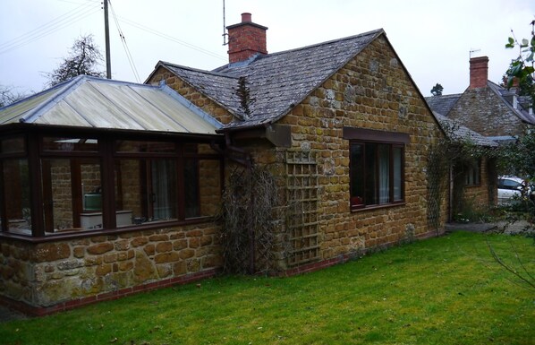 A stone house with a sloped roof, a conservatory with glass doors, and a small garden. - Copperstones, is in quiet idyllic location amid rolling hills in the Cotswolds (Banbury)