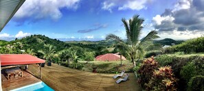 A pool area with lounge chairs and a wooden deck overlooking a hilly landscape with a red-roofed structure. - Villa Mabuya: A haven of peace with private pool and breathtaking views in Sainte-Anne (Sainte Anne)