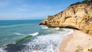 A coastal beach with clear blue water, sandy shore, and rocky cliffs.