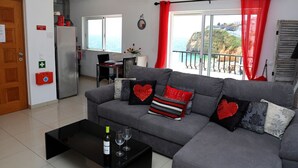 A modern living room with a grey sofa, a black coffee table with a wine bottle, and a view of the sea through large windows.