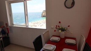 A dining area with a red table set for four, a window with a view of the beach, and a small vase with flowers on the wall.