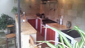 Une cuisine avec un mur recouvert de pierres, un plan de travail en bois, un évier et une cuisinière. On y voit des ustensiles de cuisine suspendus et une horloge au mur.