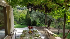 Une terrasse avec une table dressée pour le repas, entourée de chaises, et un panier de fruits posé sur la table.