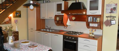 A compact kitchen with white cabinets, a gas stove, and a sink. There's a dining table with a floral tablecloth and a chair.
