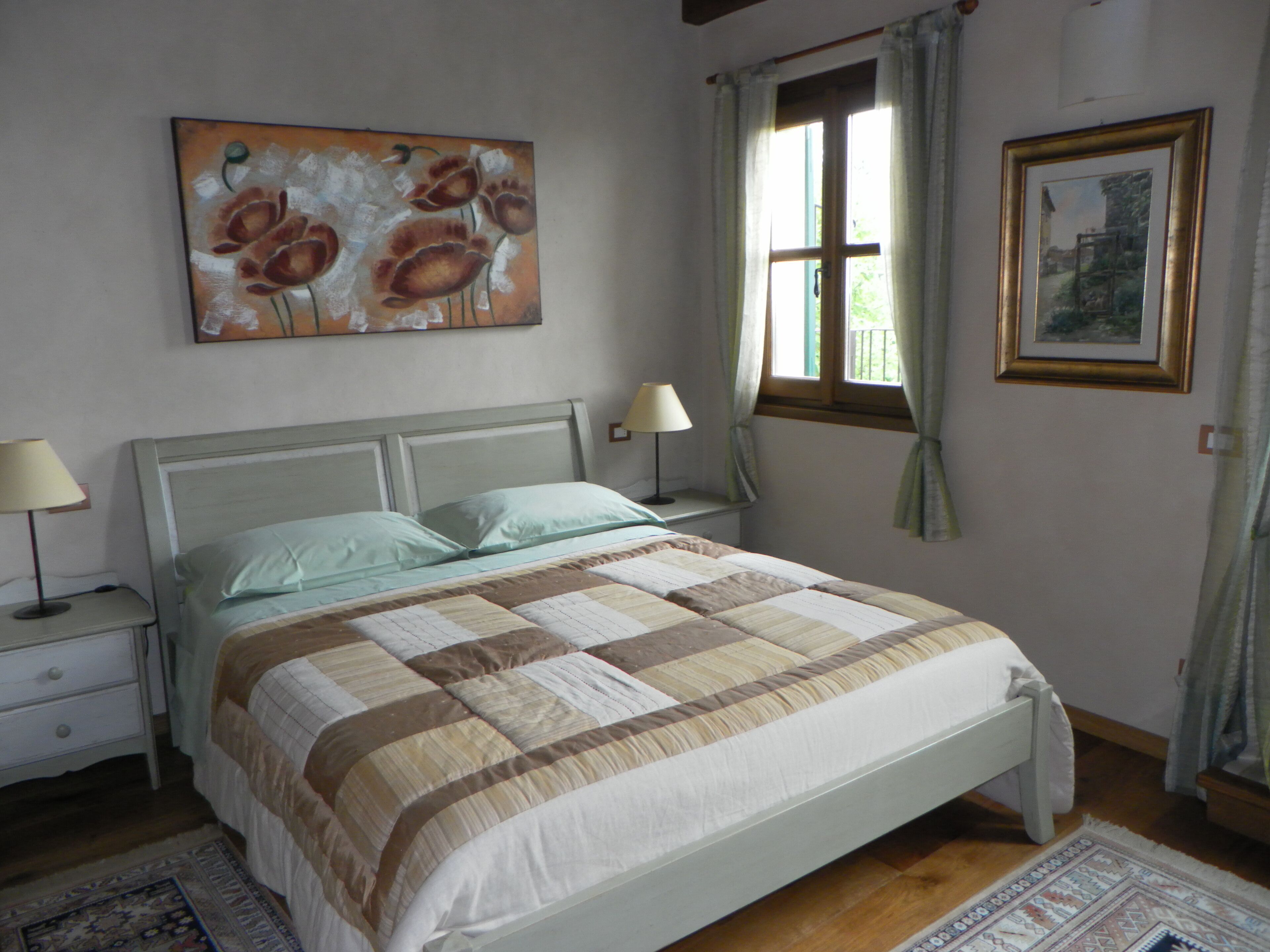 A bedroom with a bed, nightstands, a window with curtains, a painting on the wall, and a rug on the floor.