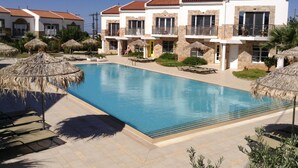 Une piscine entourée de bâtiments dotés de balcons, avec des chaises longues et des parasols, le tout sous un ciel bleu azur.