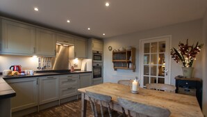 A kitchen with wooden cabinets, a central island with stools, a built-in oven, and a window with a view. - Charming Cottage in Prime Position with Castle Views, 12 The Wynding (Bamburgh)