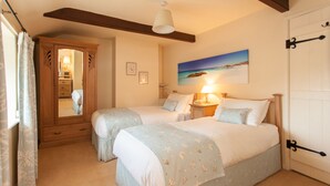 A bedroom with two beds, a wooden wardrobe, a mirror, a painting of a beach, and a door with a black handle. - Charming Cottage in Prime Position with Castle Views, 12 The Wynding (Bamburgh)