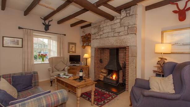 A cozy living room with a stone fireplace, two plaid sofas, a wooden coffee table, and a television. - Charming Cottage in Prime Position with Castle Views, 12 The Wynding (Bamburgh)