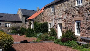 A stone building with a red-tiled roof, a small garden with various plants, and a gravel pathway. - Charming Cottage in Prime Position with Castle Views, 12 The Wynding (Bamburgh)