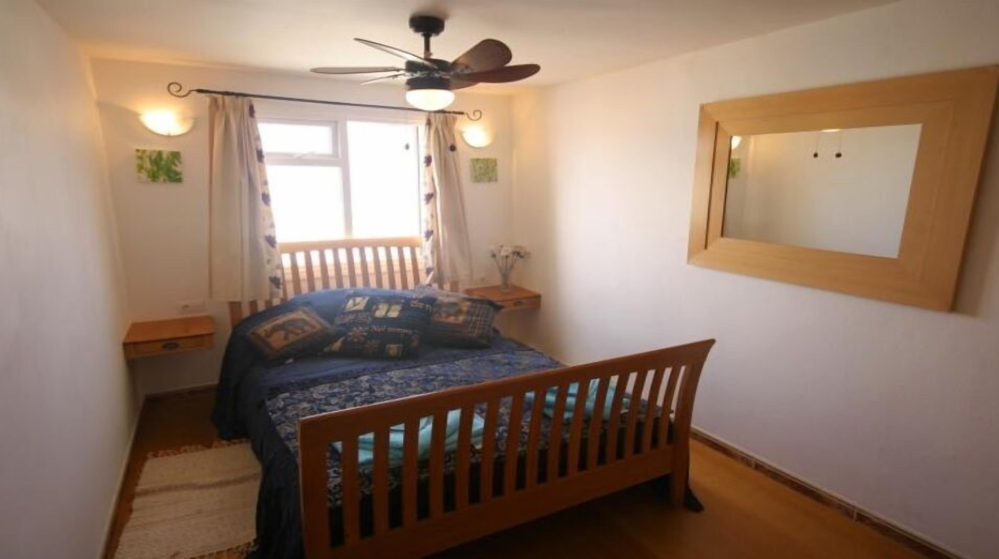 Um quarto com cama de madeira, ventilador de teto, janela com cortinas, mesa de cabeceira e espelho na parede.