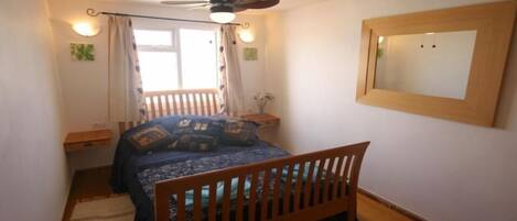 A bedroom with a wooden bed, a ceiling fan, a window with curtains, a bedside table, and a mirror on the wall.