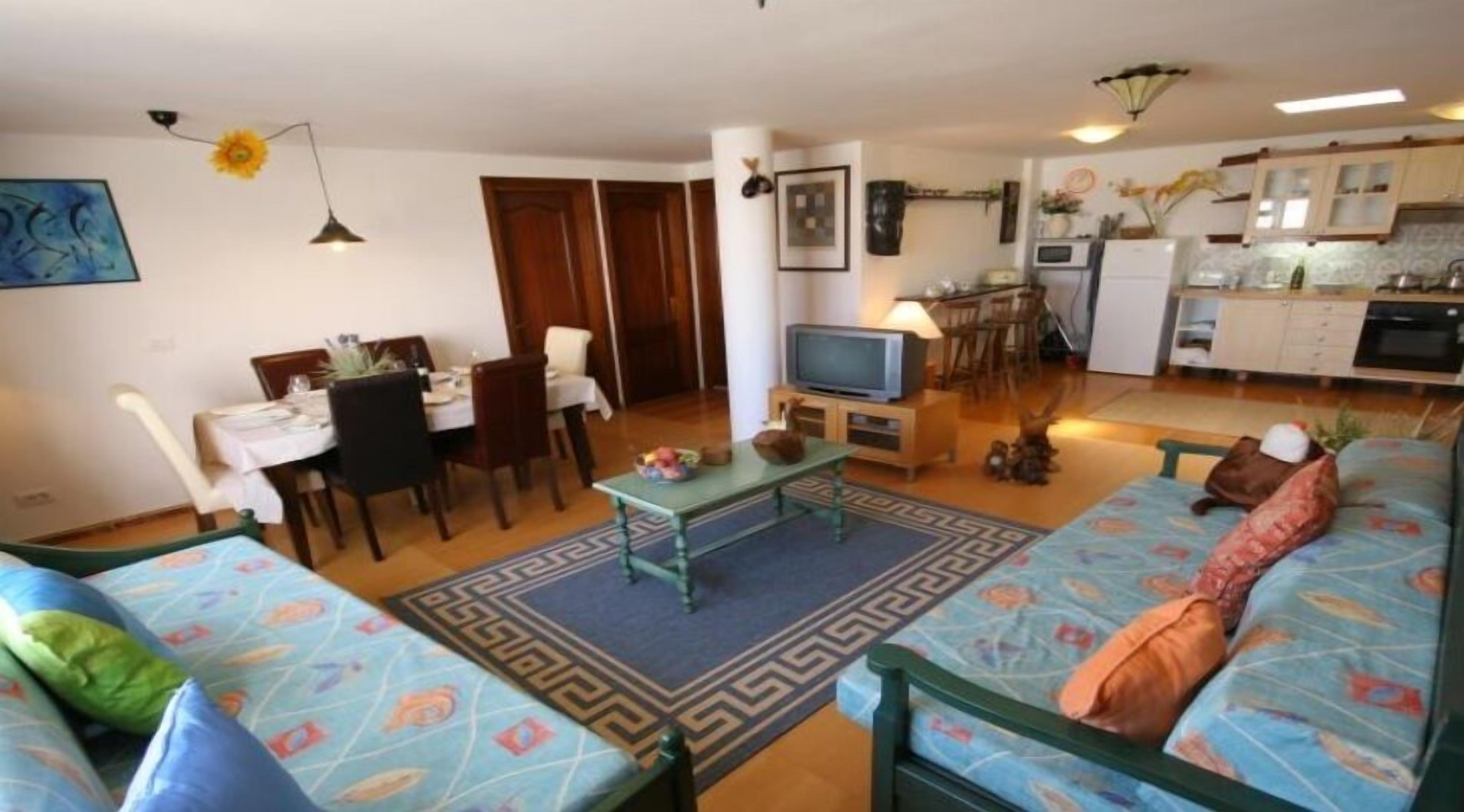 Un saló amb un sofà, una taula de dinar i cadires. Es veu una cuina al fons amb armaris i un frigorífic.