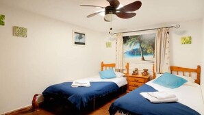 Un dormitorio con dos camas individuales, un ventilador de techo, una mesita de noche y un cuadro con temática playera en la pared.