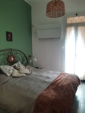 A bed with a brown blanket, a metal headboard, and decorative pillows. There is a wall-mounted air conditioner and a small table with a lamp.