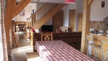 Une pièce chaleureuse avec un escalier en bois, une table à manger recouverte d’une nappe à carreaux et un coin cuisine avec un luminaire rouge suspendu.