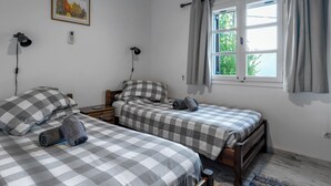 A bedroom with two beds, a window with curtains, a wall-mounted lamp, a framed picture, and a bedside table.