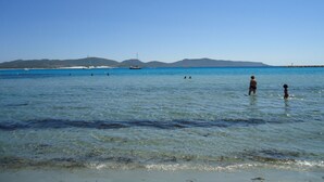 Una playa con agua azul cristalina, algunas embarcaciones y montañas a lo lejos.
