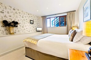 Une chambre à coucher comprenant un lit, une table de chevet, une lampe, une fenêtre avec des rideaux, un téléviseur fixé au mur et un papier peint à motifs floraux.