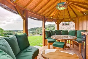 Une terrasse en bois avec une table ronde et des sièges rembourrés de couleur verte, offrant une vue sur un paysage pittoresque.